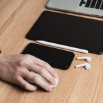 Mão usa o mouse diante de um laptop, com tablet, smartphone e fones de ouvido sobre a mesa.