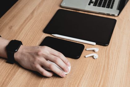 Mão usa o mouse diante de um laptop, com tablet, smartphone e fones de ouvido sobre a mesa.