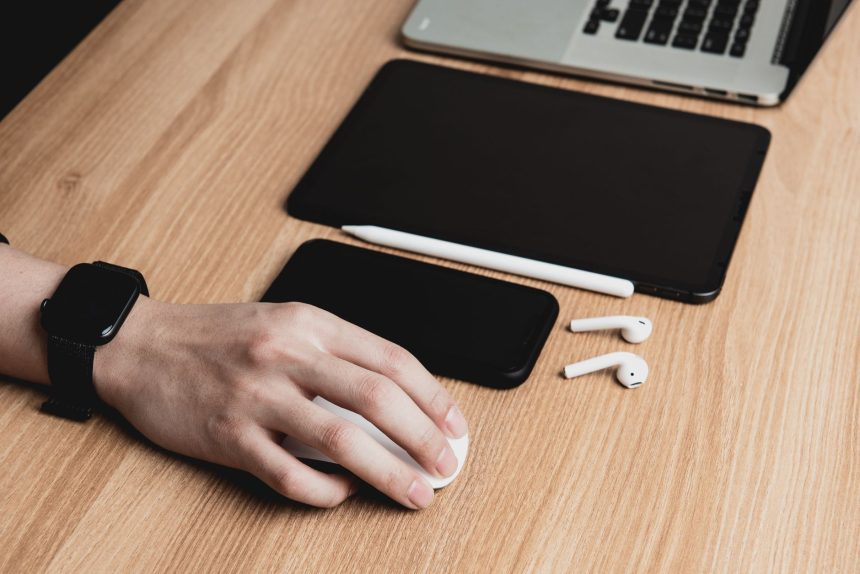 Mão usa o mouse diante de um laptop, com tablet, smartphone e fones de ouvido sobre a mesa.