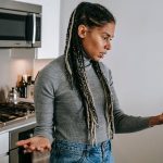 Mulher com tranças conversa por videochamada pelo celular, com expressão séria e gestos de desagrado em uma cozinha.