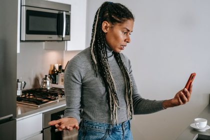 Mulher com tranças conversa por videochamada pelo celular, com expressão séria e gestos de desagrado em uma cozinha.