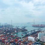 Vista aérea de um movimentado terminal portuário de contêineres, com navios, guindastes e áreas industriais.