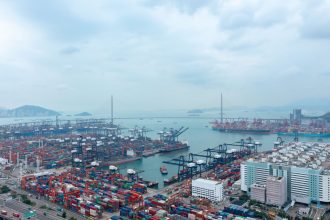 Vista aérea de um movimentado terminal portuário de contêineres, com navios, guindastes e áreas industriais.
